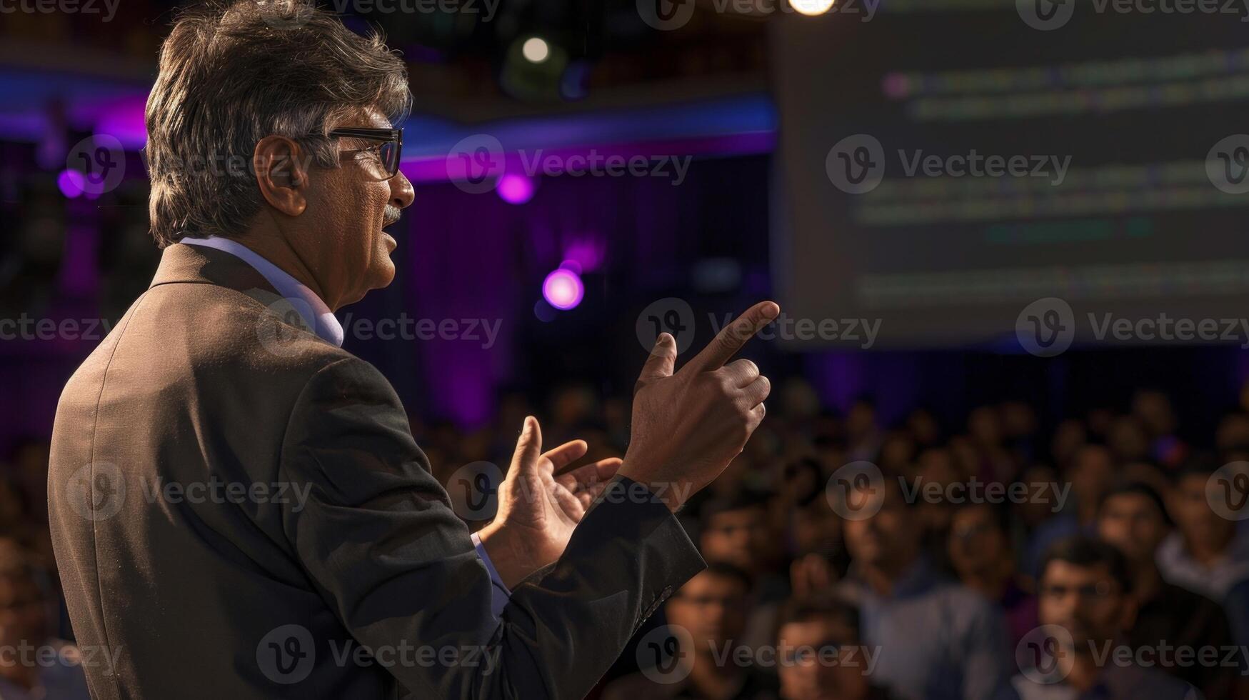 A middle aged Indian man in business attire and glasses gives an inspiring speech to a large audience photo