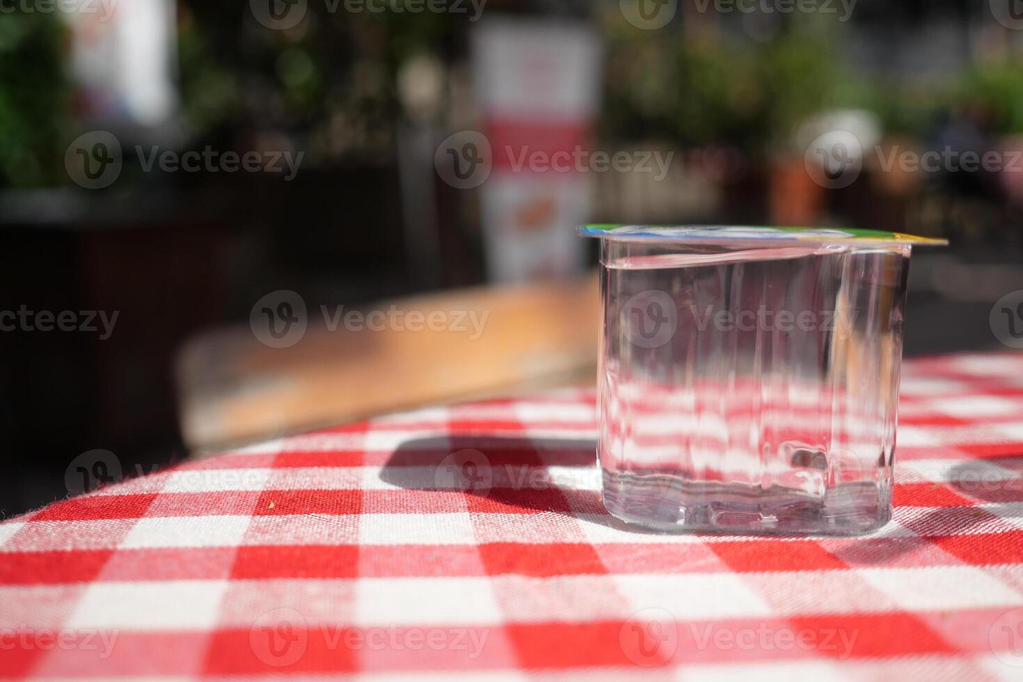 water in a plastic cup on a checkered table outside, a glass cup on a red and white tablecloth photo