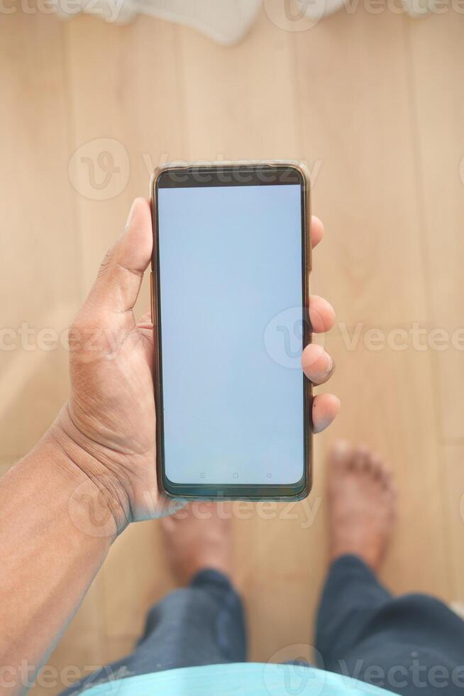 A hand is holding a smartphone with a blank screen on a wooden floor in a closeup shot photo