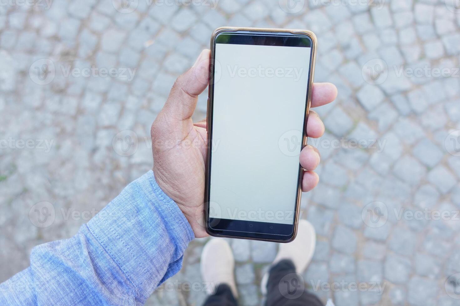 Person in blue shirt holds smartphone with blank screen, outdoors on cobblestone background photo
