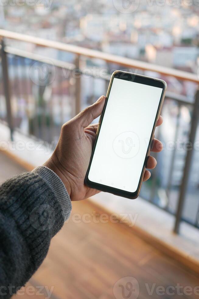 A hand holding a smartphone with a blank screen on a balcony with a city view in the background photo