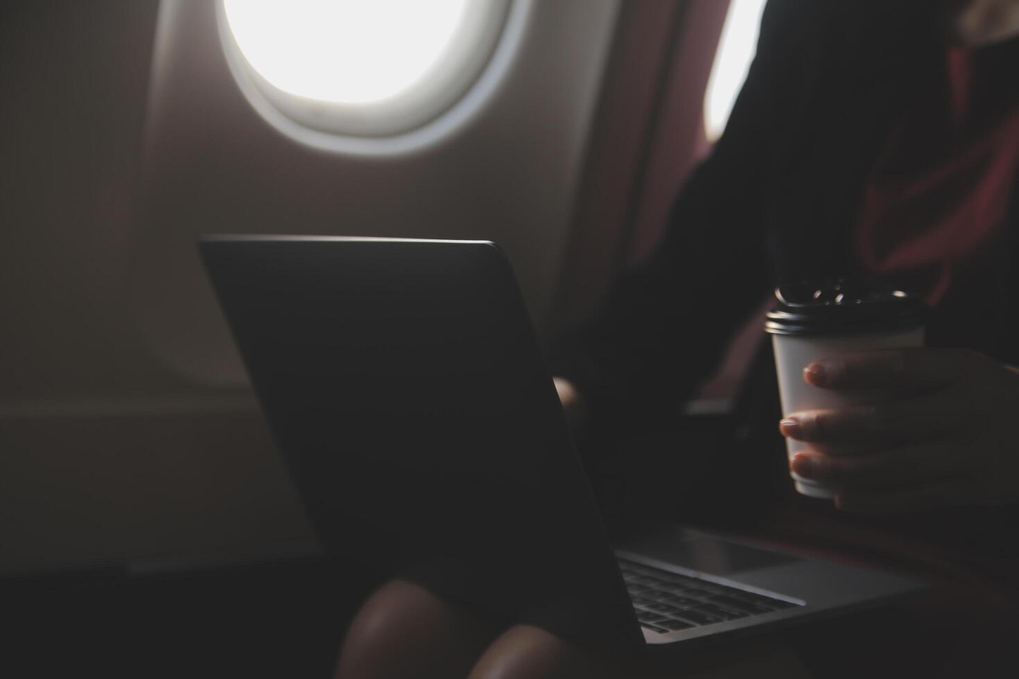 Blonde female tourist checking incoming notification on smartphone sitting on seat of airplane with netbook.Young businesswoman share media from telephone on laptop computer during plane flight photo