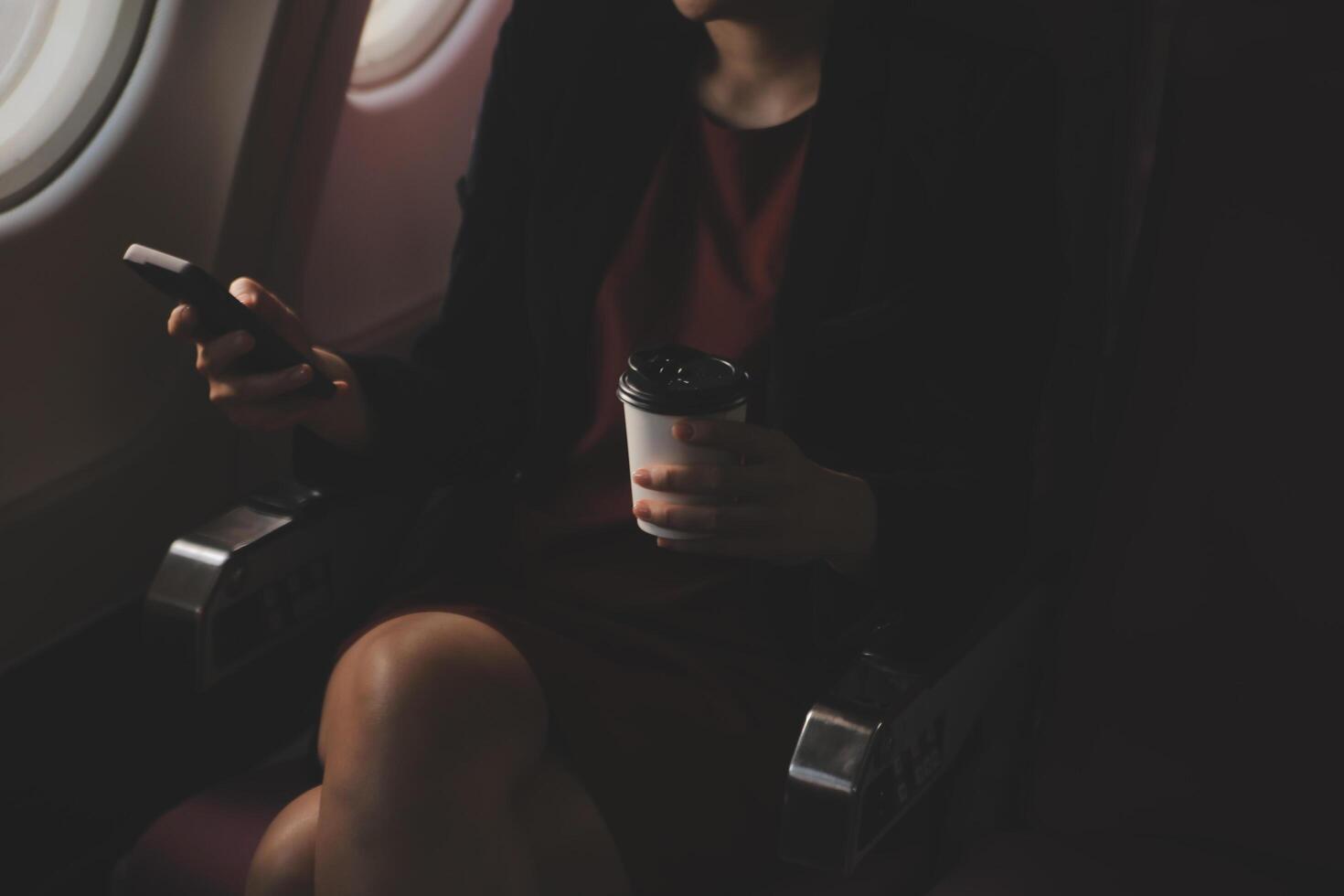 Blonde female tourist checking incoming notification on smartphone sitting on seat of airplane with netbook.Young businesswoman share media from telephone on laptop computer during plane flight photo