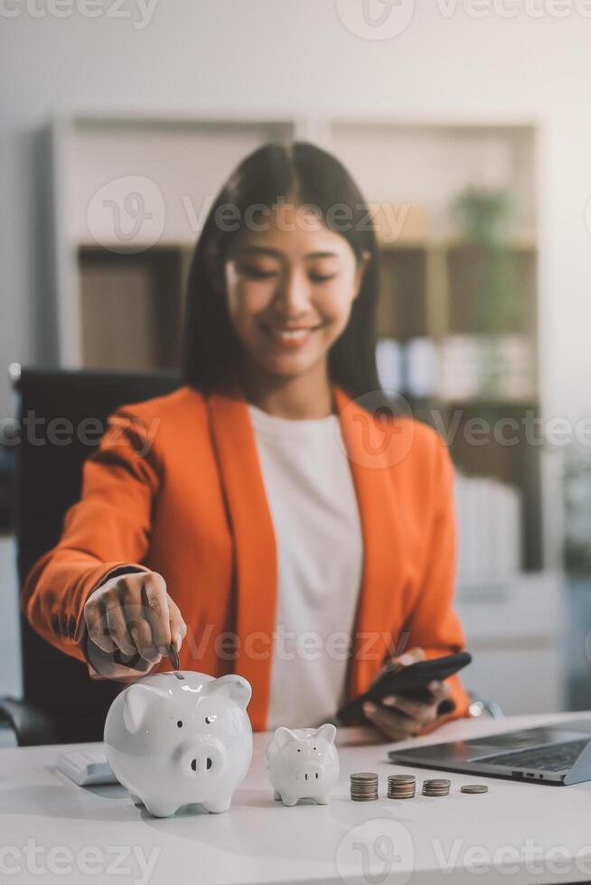 Beautiful Asian businesswoman insert coins in a pin piggy bank on the table. saving money, investment, currency concept photo