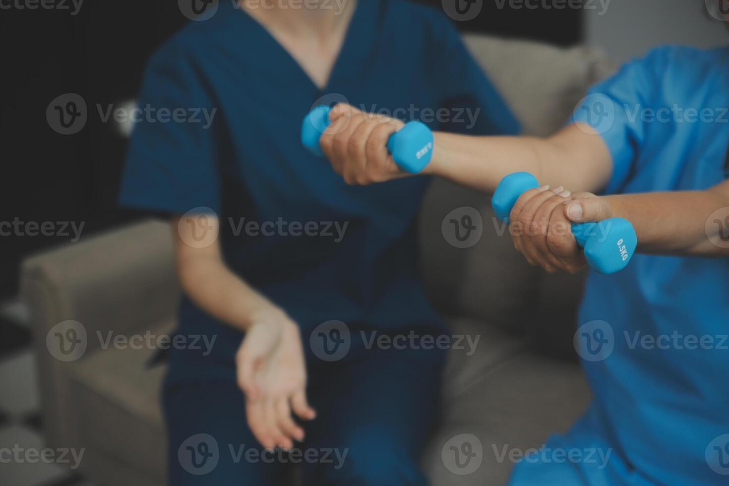 Old woman training with physiotherapist using dumbbells at home. Therapist assisting senior woman with exercises in nursing home. Elderly patient using dumbbells with outstretched arms. photo