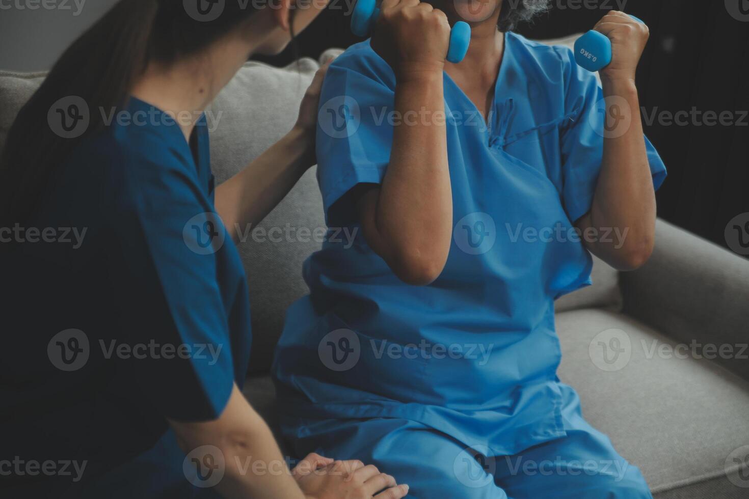 Old woman training with physiotherapist using dumbbells at home. Therapist assisting senior woman with exercises in nursing home. Elderly patient using dumbbells with outstretched arms. photo