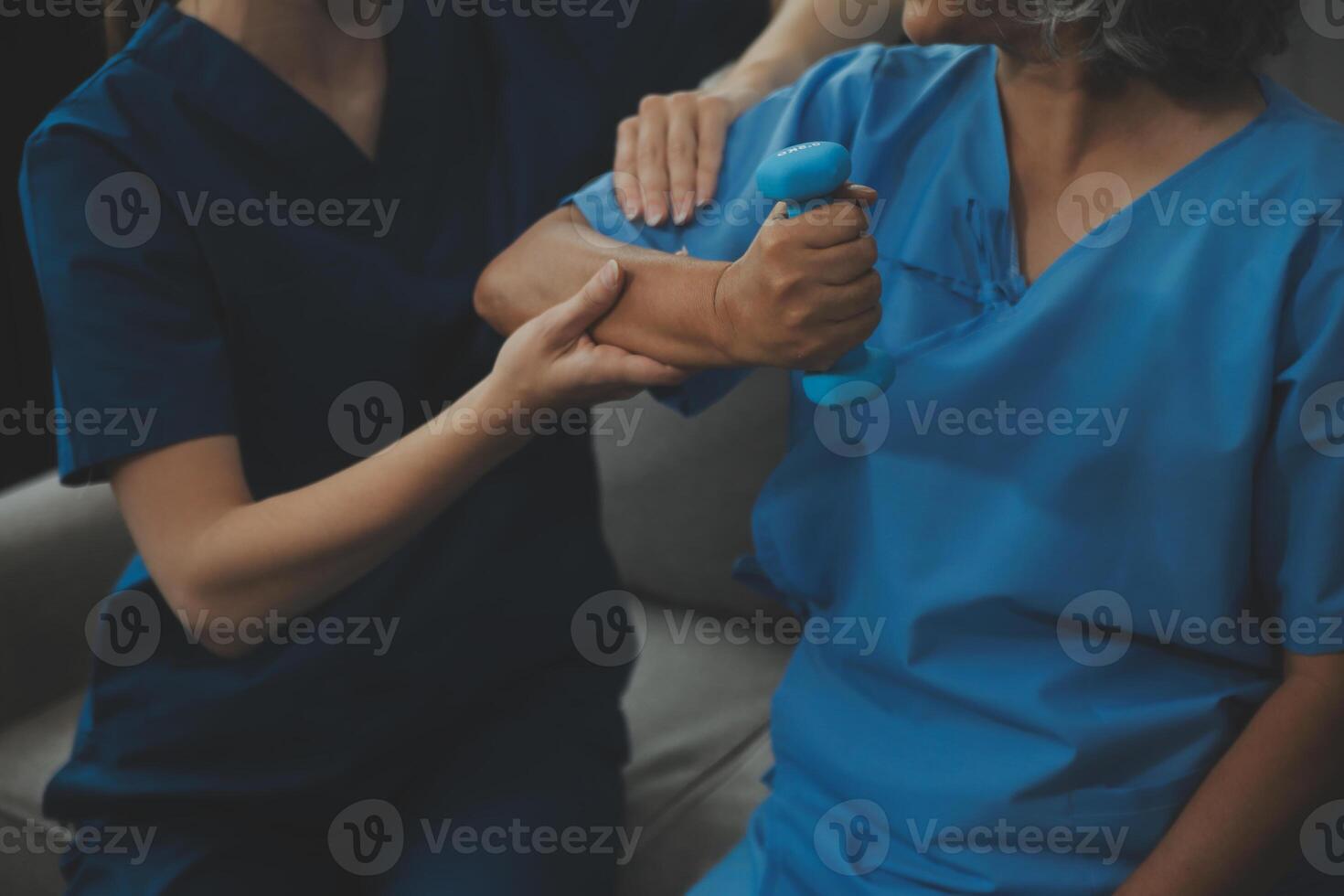 Old woman training with physiotherapist using dumbbells at home. Therapist assisting senior woman with exercises in nursing home. Elderly patient using dumbbells with outstretched arms. photo