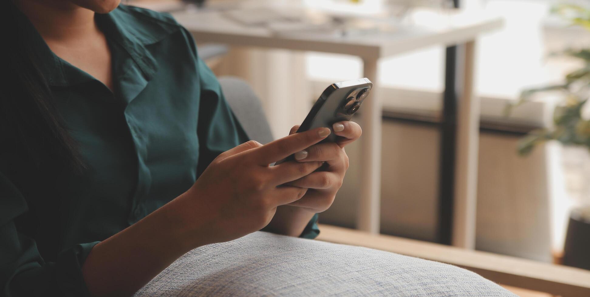 Charming young Asian woman using cellphone, communicating on web, working or learning online, sitting in armchair against white wall, free space. Lovely millennial lady chatting on smartphone photo