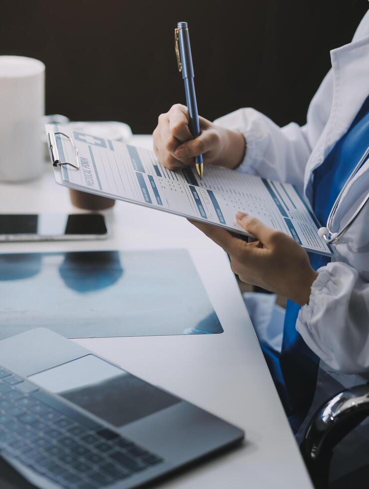 A beautiful and experienced young Asian female doctor in a white gown is working at her desk in the hospital, reading medical reports online. photo