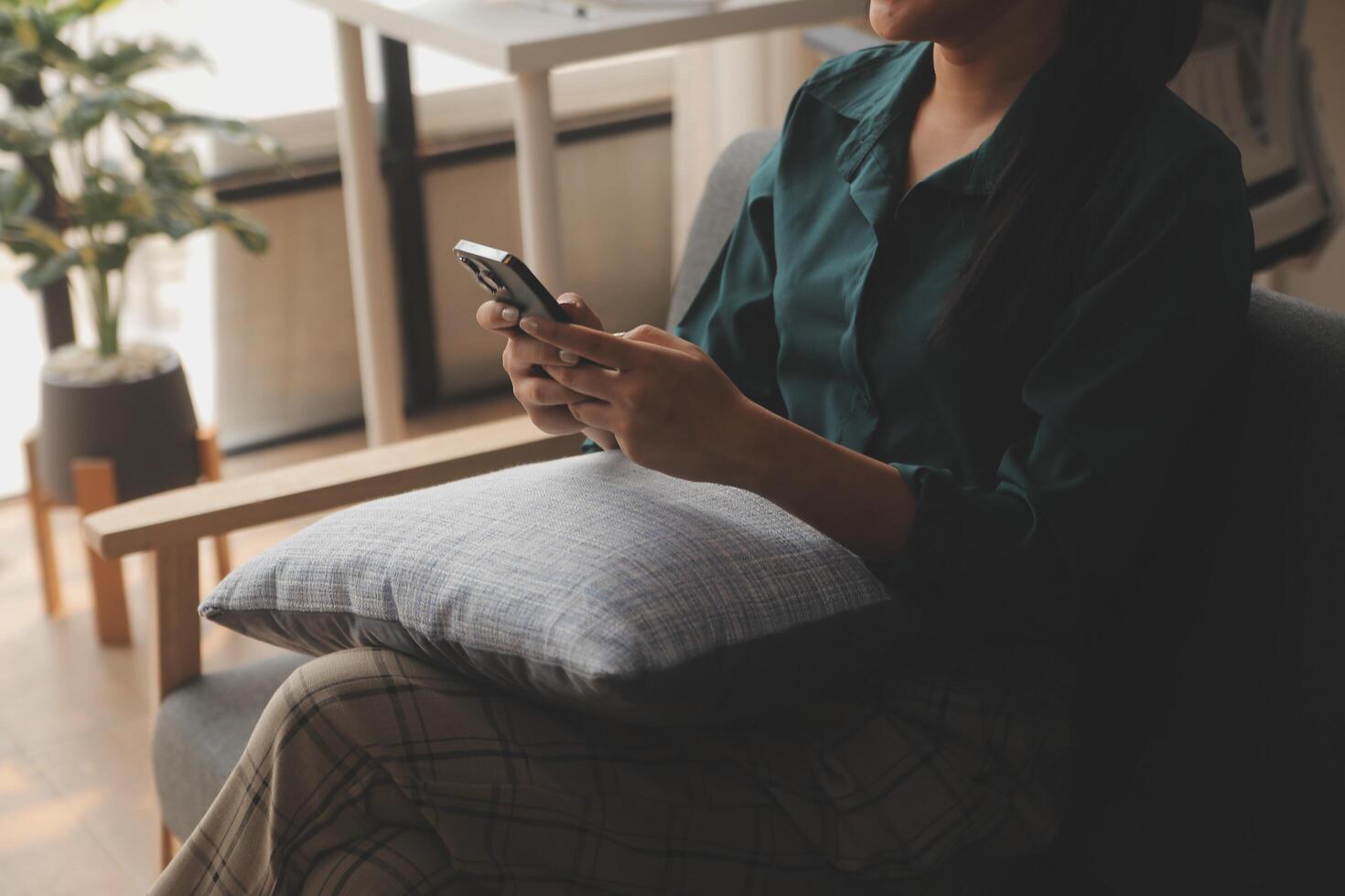 Charming young Asian woman using cellphone, communicating on web, working or learning online, sitting in armchair against white wall, free space. Lovely millennial lady chatting on smartphone photo