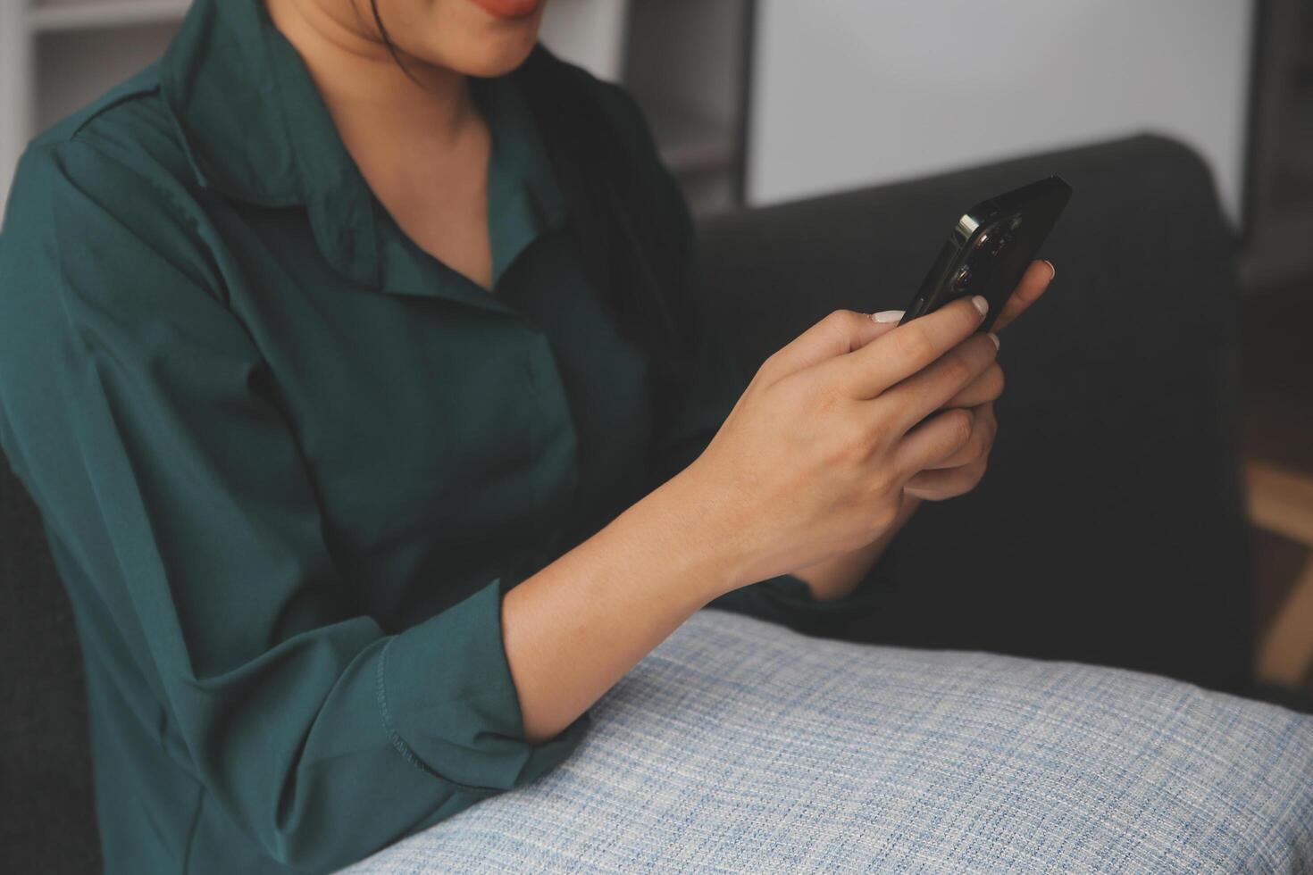 Charming young Asian woman using cellphone, communicating on web, working or learning online, sitting in armchair against white wall, free space. Lovely millennial lady chatting on smartphone photo