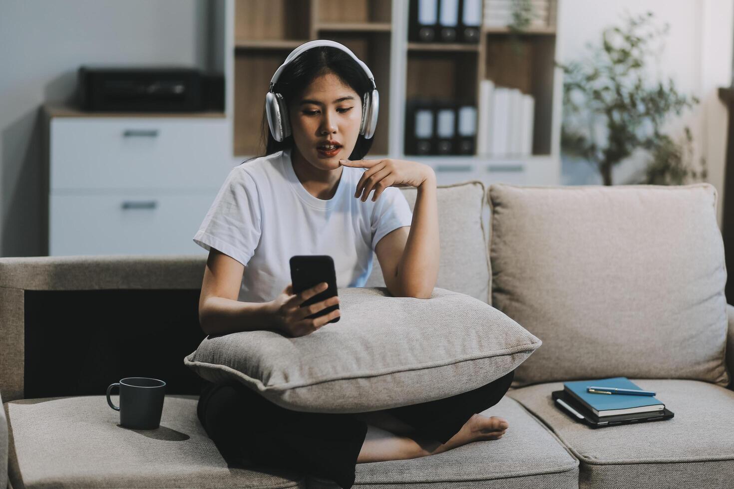 Young asian woman using smartphone and wearing headphone sitting on couch at home. Happy girl relaxing comfortable in the living room, spending lazy weekend, listening to music, watching movie. photo