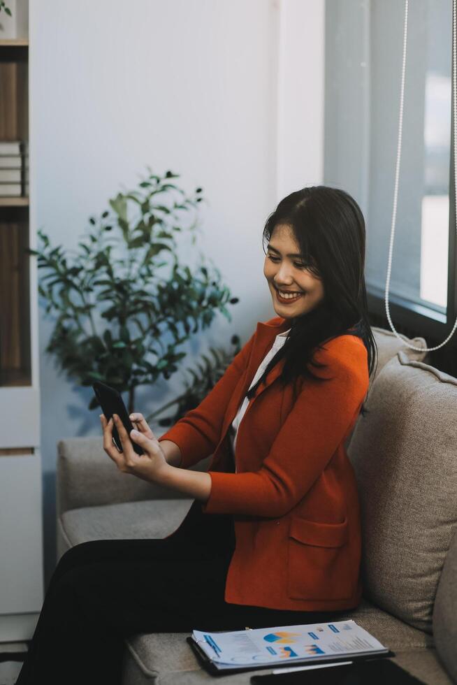 Smiling and happy millennial Asian businesswoman chatting with her friends on her phone, scrolling on social media or watching on the internet, using her phone while relaxing in the office. photo