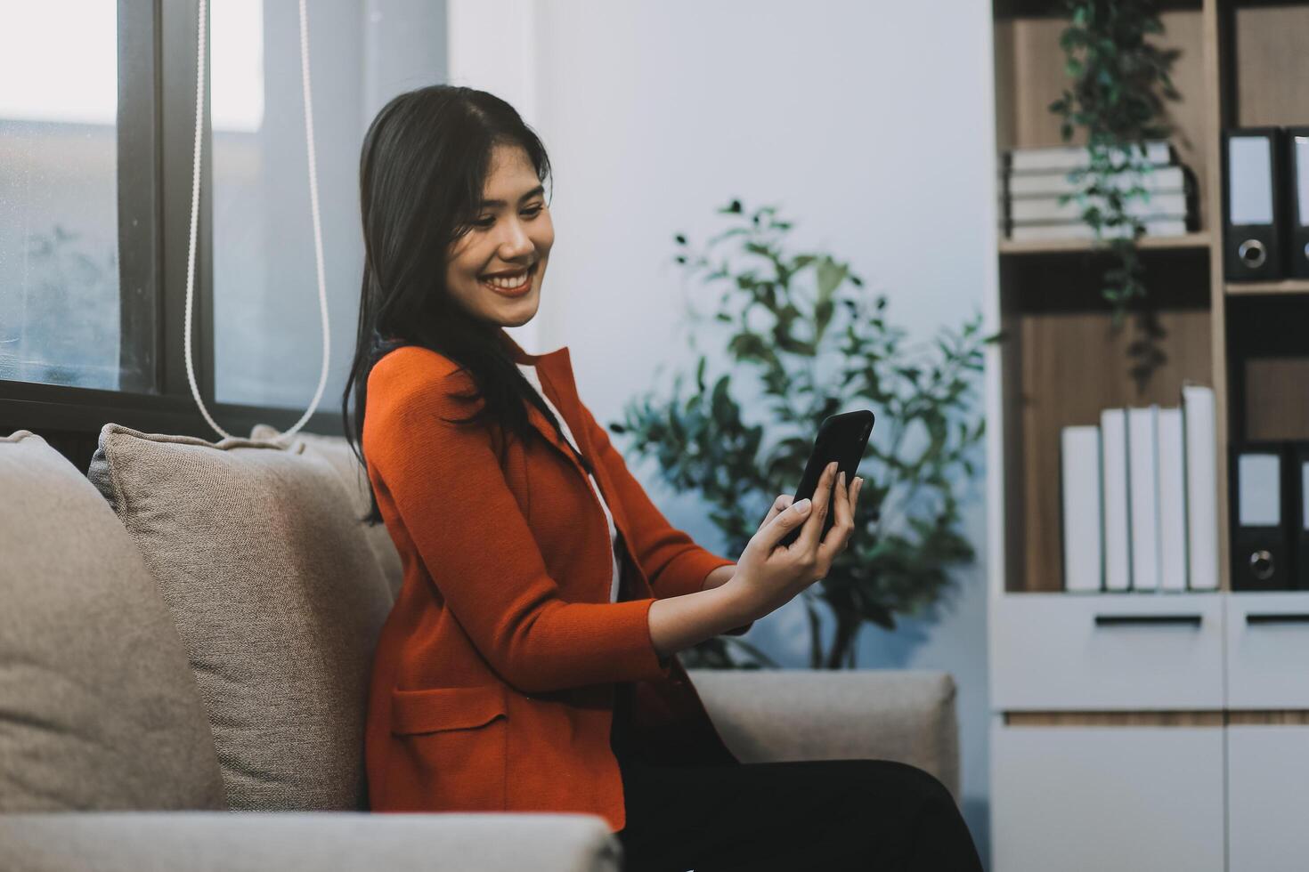 Smiling and happy millennial Asian businesswoman chatting with her friends on her phone, scrolling on social media or watching on the internet, using her phone while relaxing in the office. photo