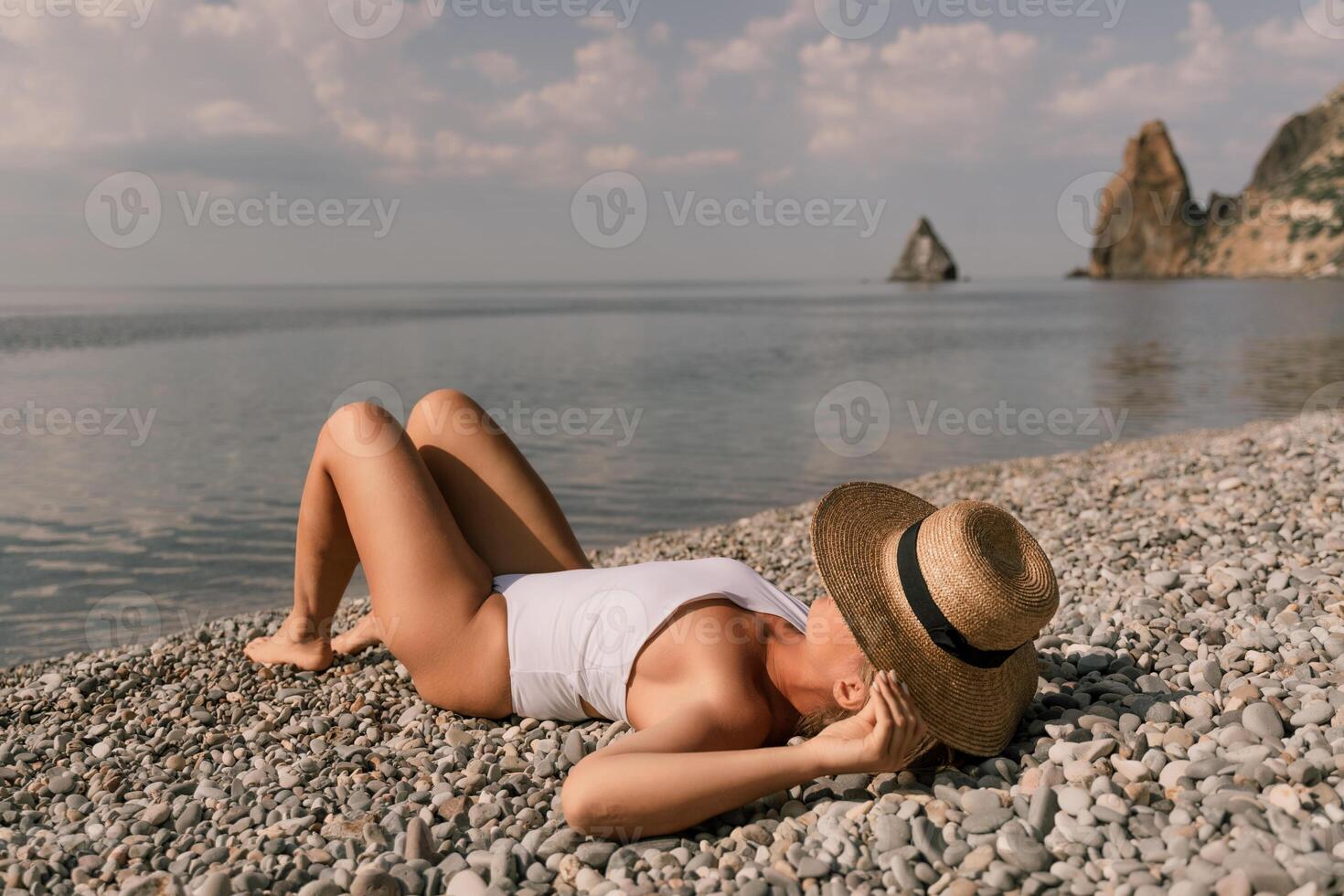 A woman is laying on the beach wearing a straw hat. She is wearing a white bikini top and white bikini bottoms. photo