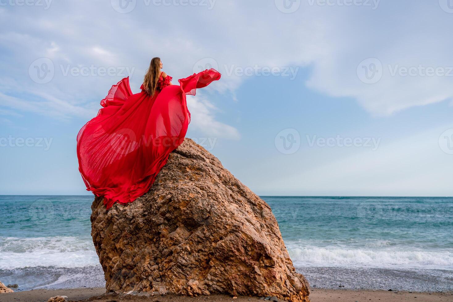 woman sea red dress. Woman with long hair on a sunny seashore in a red flowing dress, back view, silk fabric waving in the wind. Against the backdrop of the blue sky and mountains on the seashore. photo