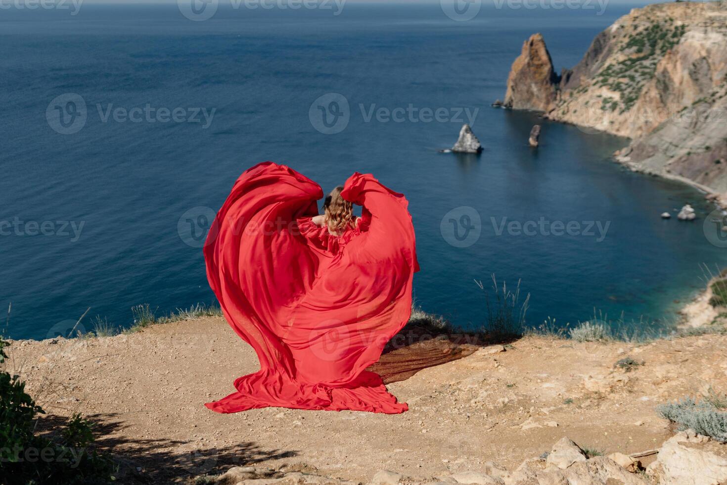 A woman in a red dress is standing on a rocky beach, with the ocean in the background. The woman is wearing a red dress that is flowing out in a heart shape, giving the impression of a romantic photo