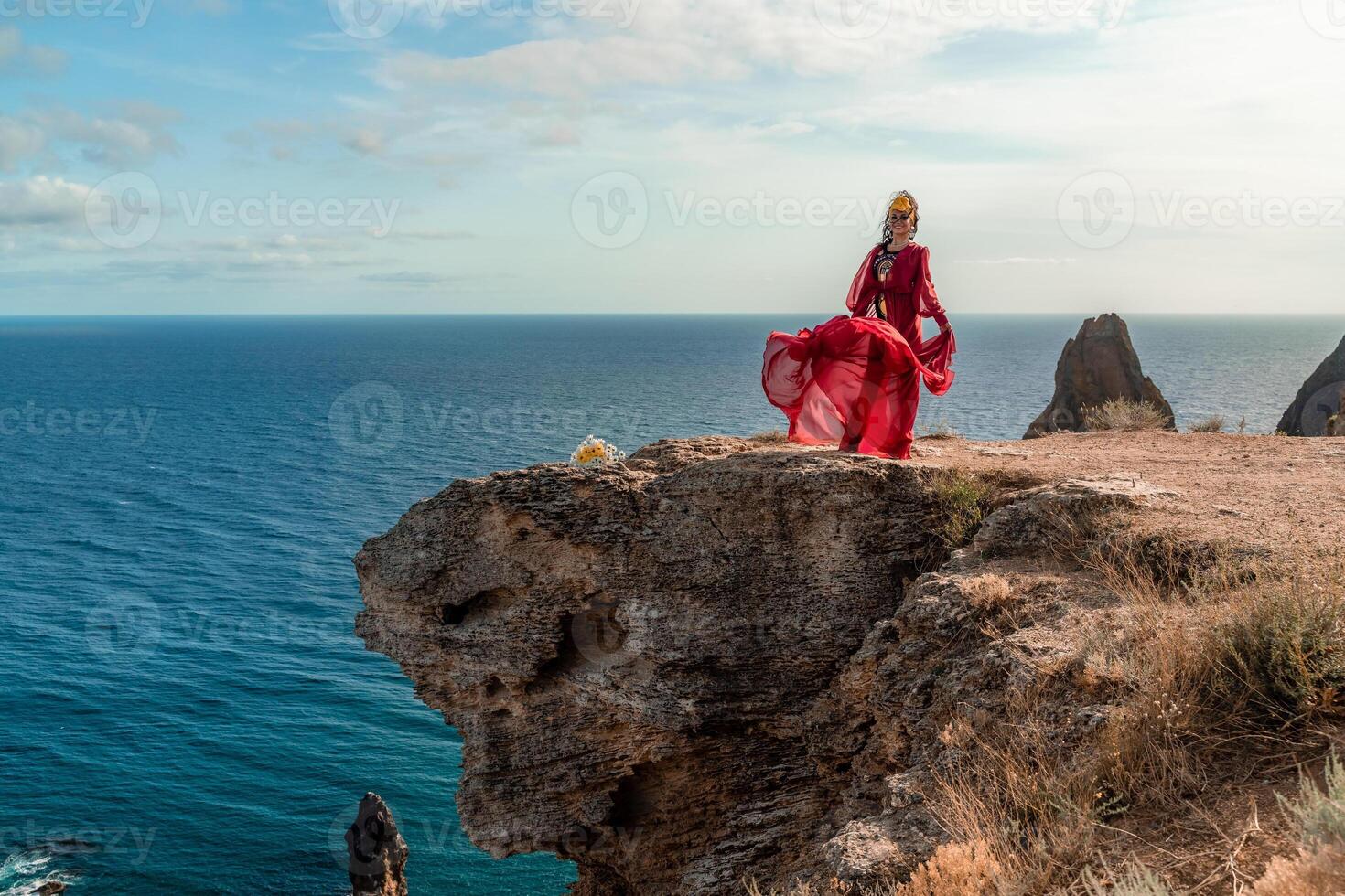 A woman in a red dress stands on a rocky cliff overlooking the ocean. The scene is serene and peaceful, with the woman's flowing dress adding a sense of grace and beauty to the landscape. photo