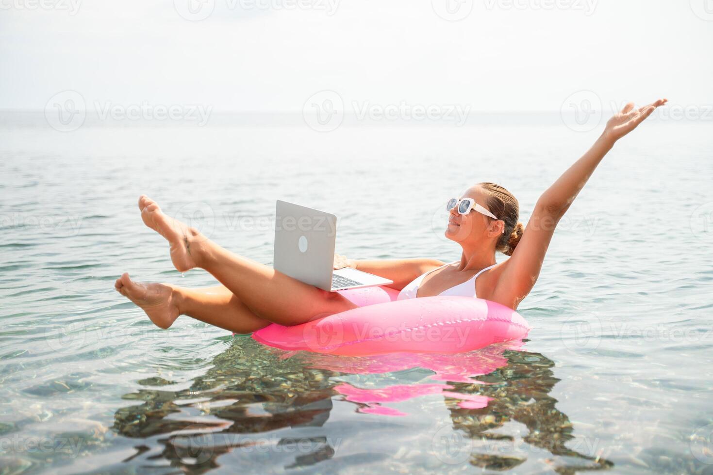 A woman is floating on a pink inflatable raft with a laptop on her lap. She is wearing sunglasses and she is enjoying the sun and the water. Concept of relaxation and leisure. photo