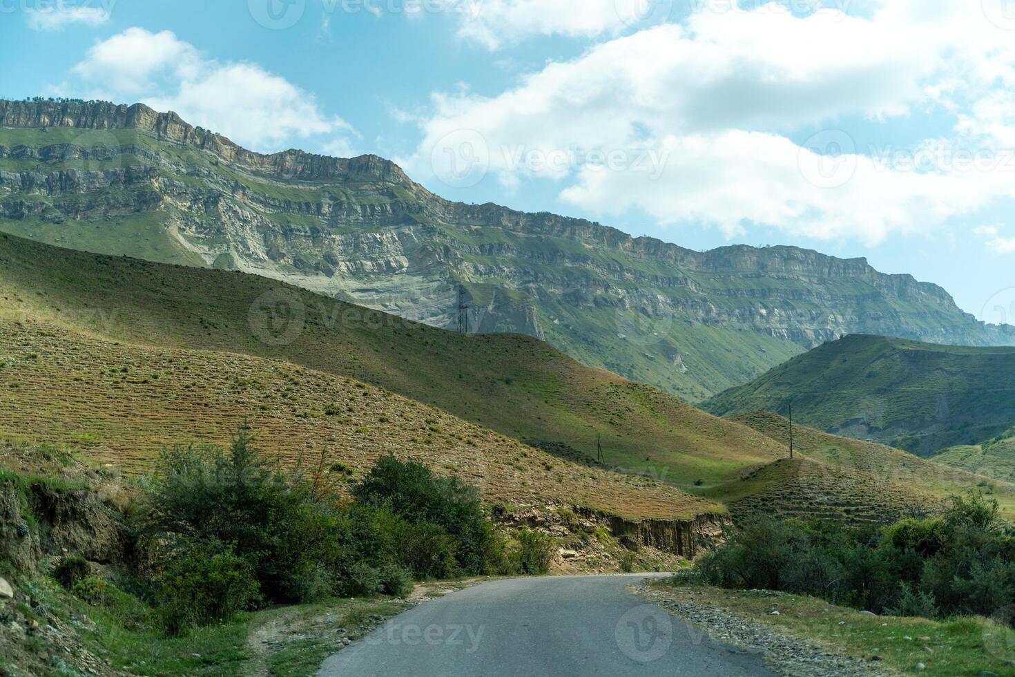 A road runs through a valley with mountains in the background. The road is surrounded by trees and grass, and the sky is clear and blue. photo