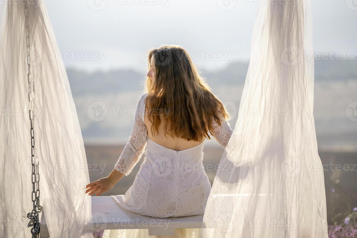 A woman in a white dress sits on a bench under a white canopy. The scene is serene and peaceful, with the woman looking out over the landscape. photo