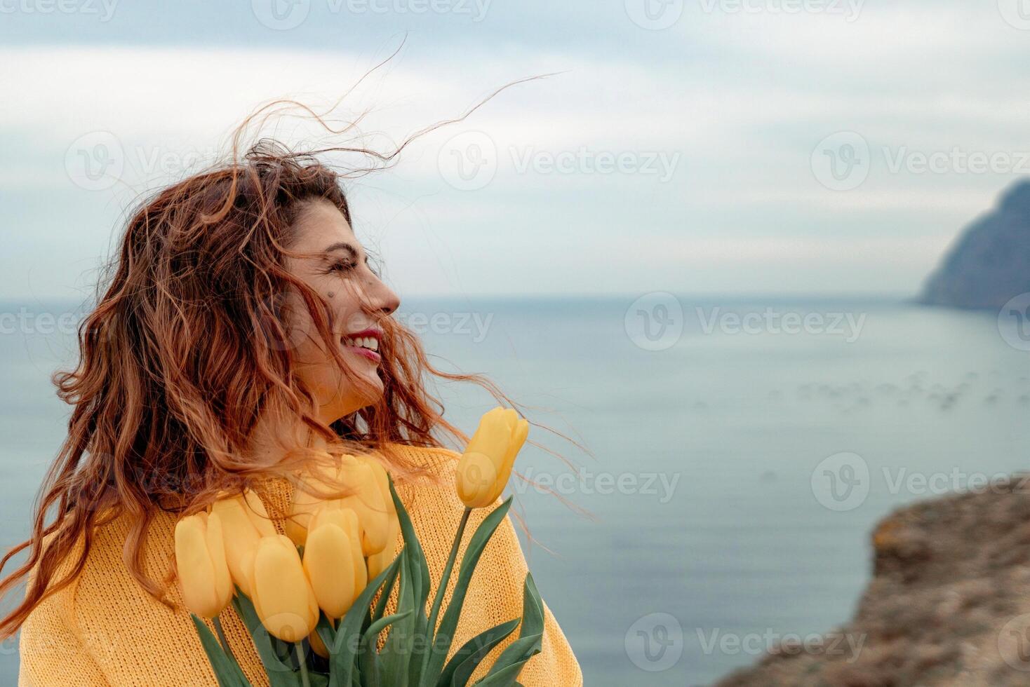 Portrait of a happy woman with hair flying in the wind against the backdrop of mountains and sea. Holding a bouquet of yellow tulips in her hands, wearing a yellow sweater photo