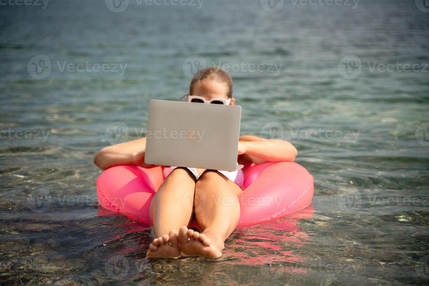 A woman is sitting on a pink inflatable raft in the ocean, using a laptop. Concept of relaxation and leisure, as the woman is enjoying her time in the water while working on her laptop. photo