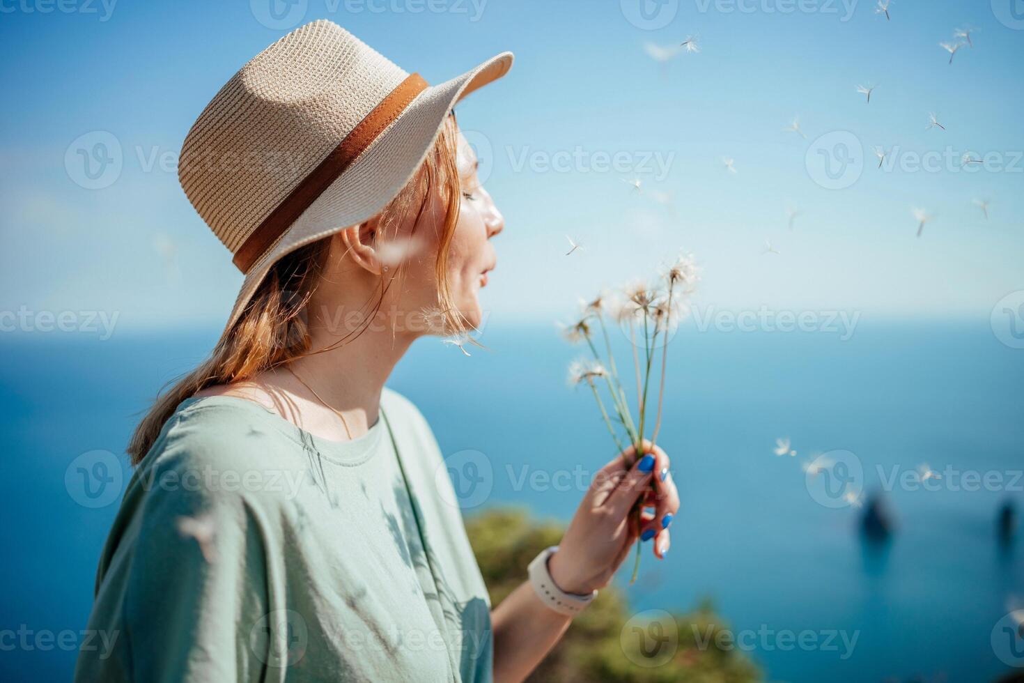 A woman wearing a straw hat is blowing on a bunch of dandelions. The scene is set on a beach with a blue ocean in the background. The woman is enjoying the moment and the beauty of the flowers. photo