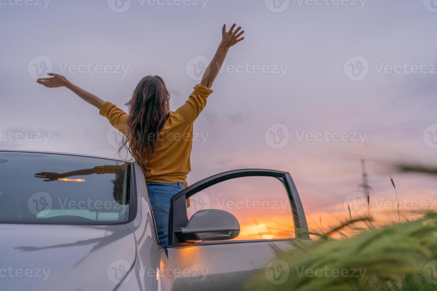 A woman is standing in the open door of a car, with her arms raised in the air photo