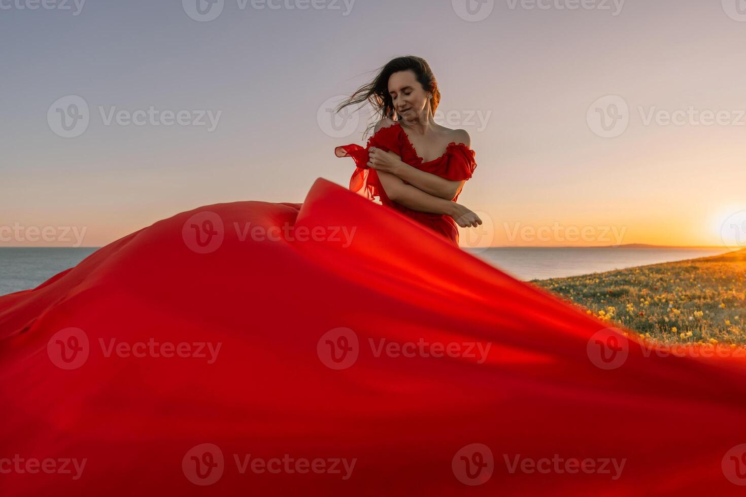 woman red dress standing grassy hillside. The sun is setting in the background, casting a warm glow over the scene. The woman is enjoying the beautiful view and the peaceful atmosphere. photo