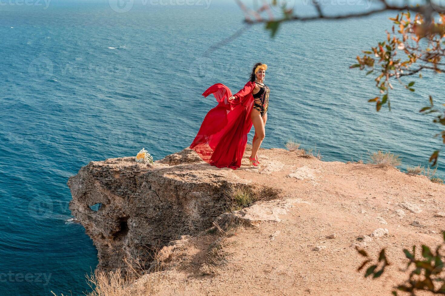 A woman in a red dress is standing on a rocky cliff overlooking the ocean. She is wearing a lei and she is dancing or celebrating. The scene is serene and peaceful, with the ocean as a backdrop. photo