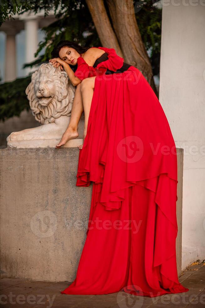 A woman in a red dress is sitting on a stone lion statue. The statue is white and has a lion's head. The woman is wearing a black belt and is looking at the camera. The scene has a calm. photo