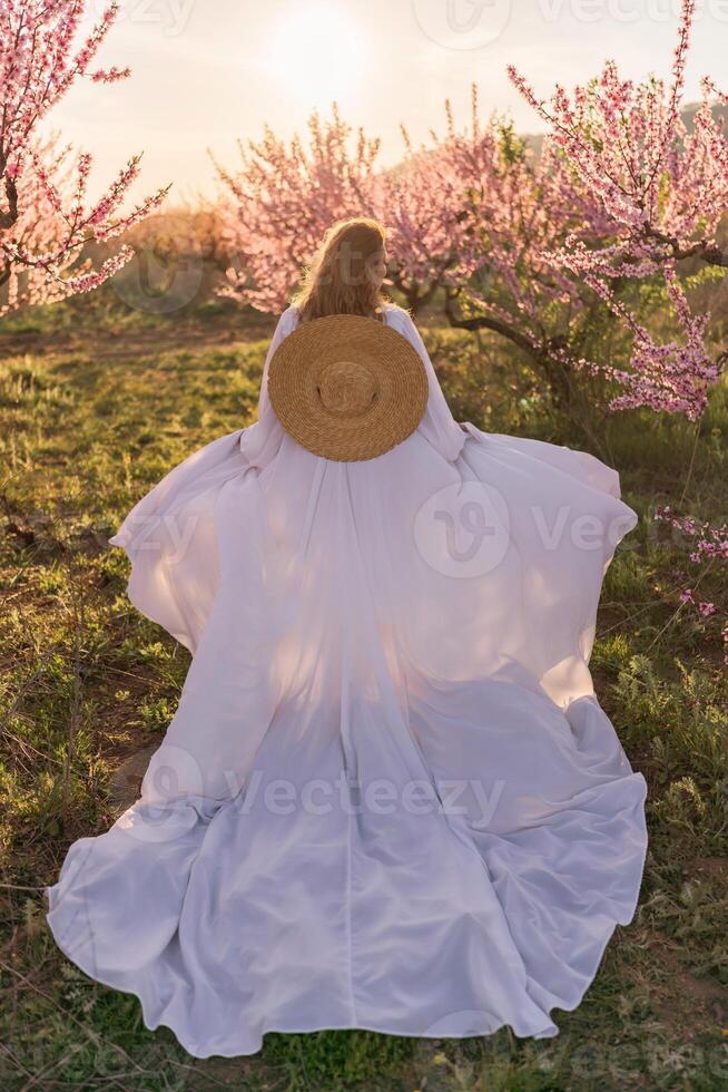 Woman blooming peach orchard. Against the backdrop of a picturesque peach orchard, a woman in a long white dress and hat enjoys a peaceful walk in the park, surrounded by the beauty of nature. photo