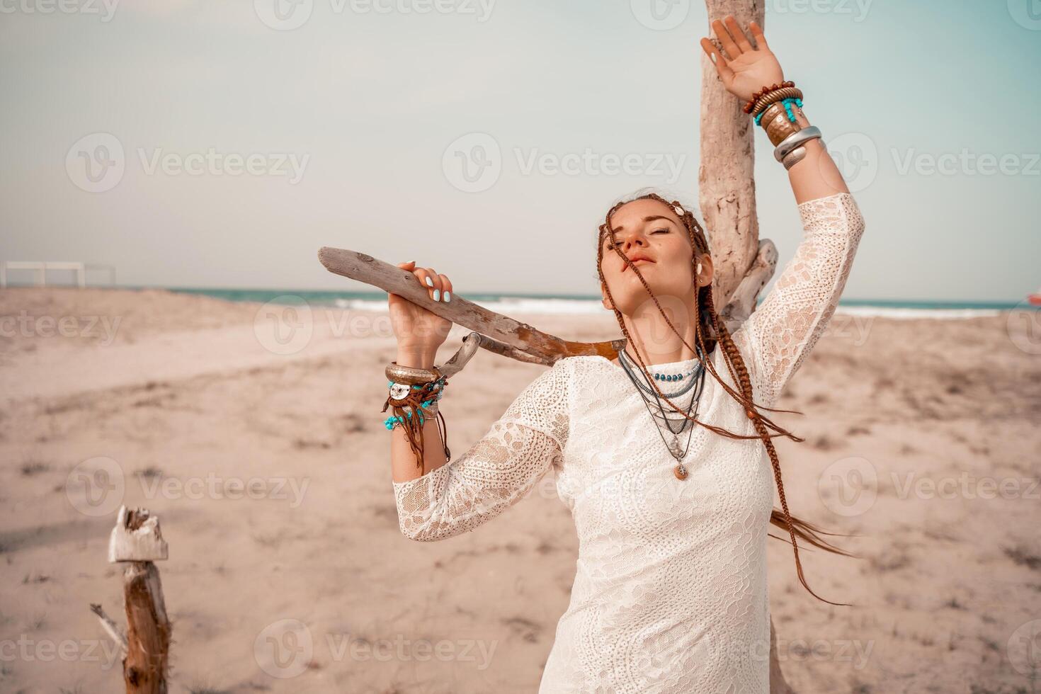 Model in boho style in a white long dress and silver jewelry on the beach. Her hair is braided, and there are many bracelets on her arms. photo