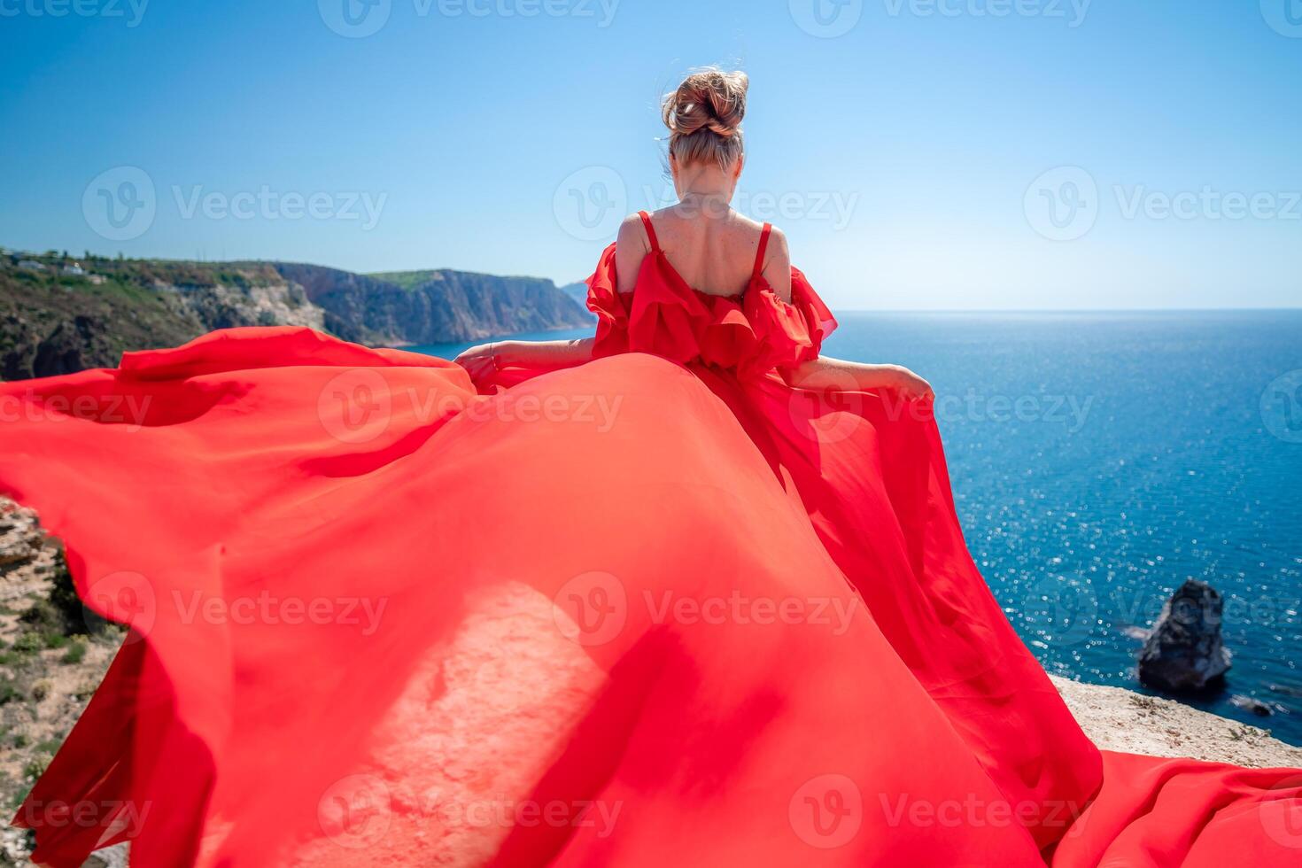 Blonde with long hair on a sunny seashore in a red flowing dress, back view, silk fabric waving in the wind. Against the backdrop of the blue sky and mountains on the seashore. photo