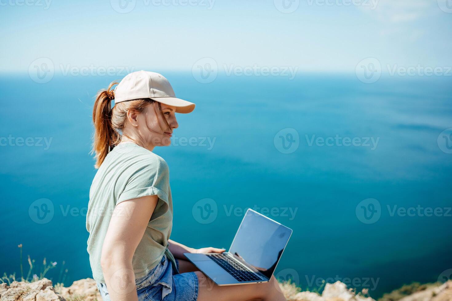 A woman is sitting on a rock by the ocean with a laptop in front of her. She is wearing a hat and has her hair in a ponytail. Concept of relaxation and leisure. photo