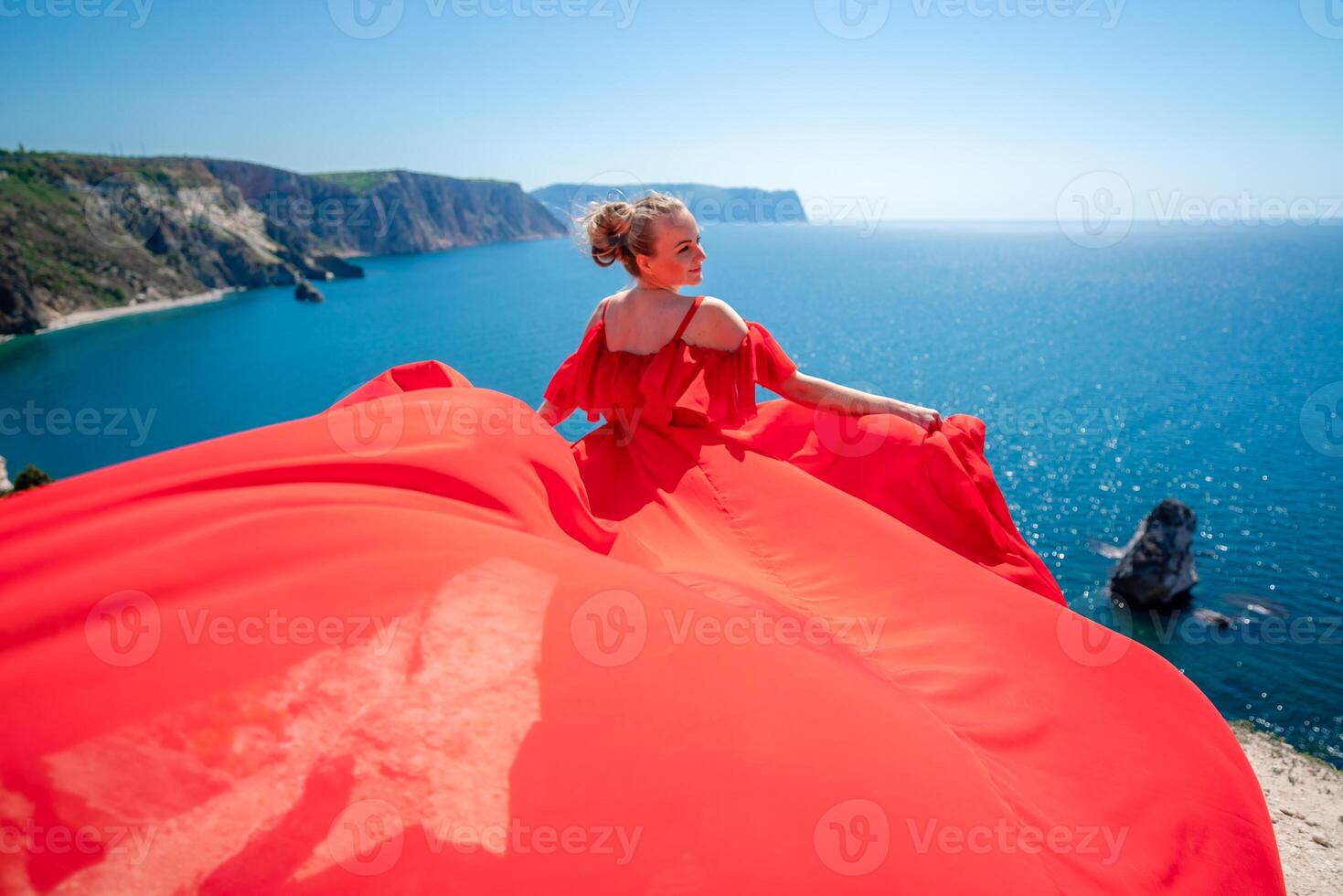 Blonde with long hair on a sunny seashore in a red flowing dress, back view, silk fabric waving in the wind. Against the backdrop of the blue sky and mountains on the seashore. photo