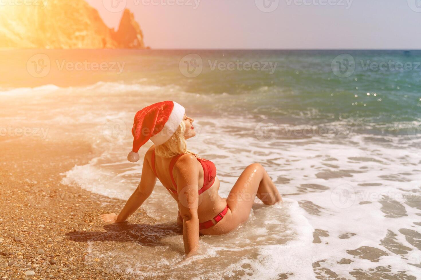 Female beach Santa hat wave coast. beach relaxation seaside. A woman in a red swimsuit enjoying her time on the beach, sits on the sand and being covered by a wave. photo