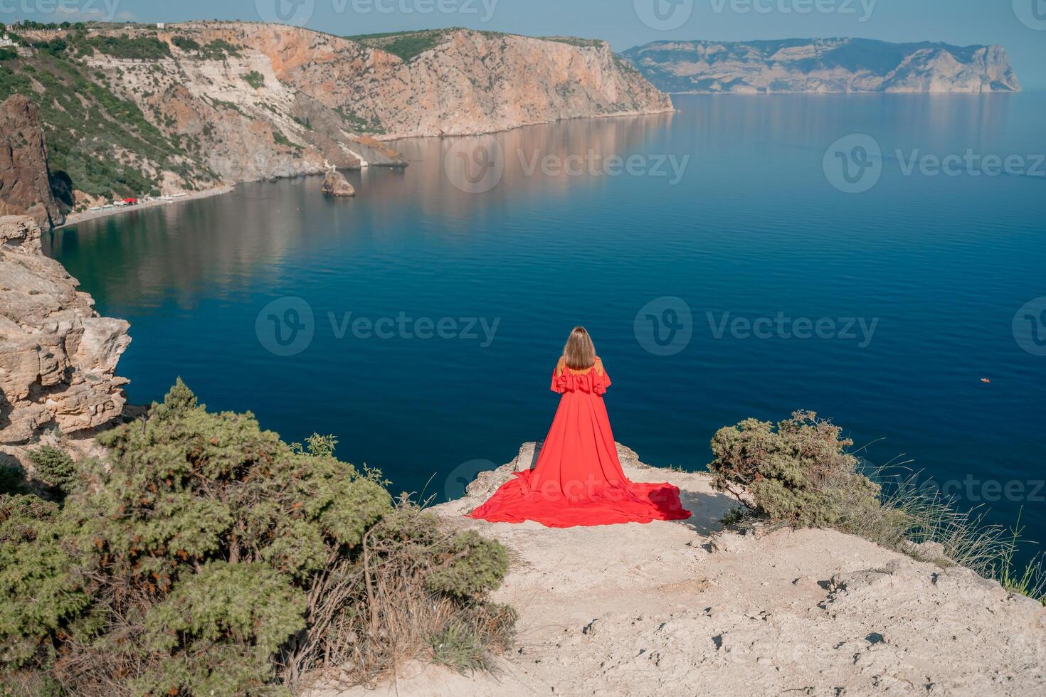 Red dress sea woman. Happy woman with flowing hair in a long flowing red dress stands on a rock near the sea. Travel concept, photo session at sea