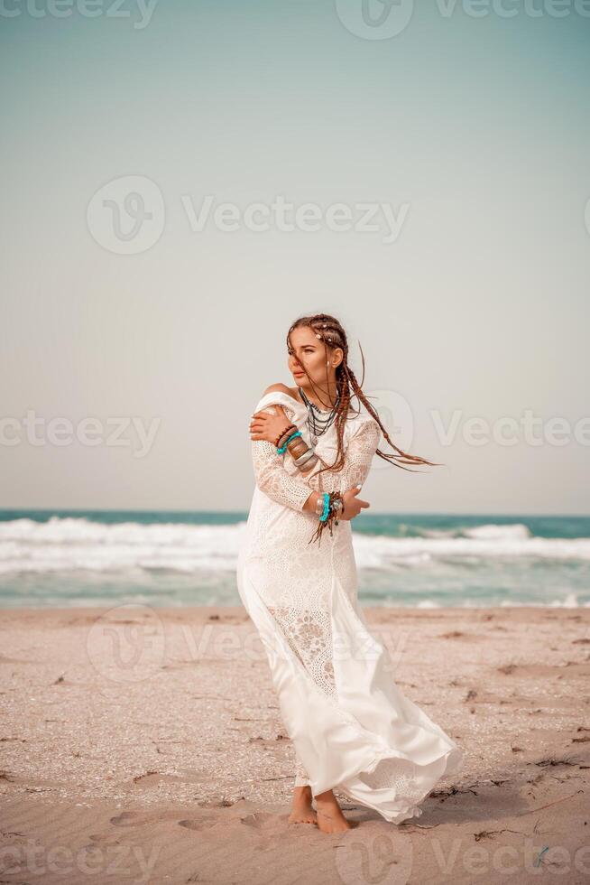 Model in boho style in a white long dress and silver jewelry on the beach. Her hair is braided, and there are many bracelets on her arms. photo