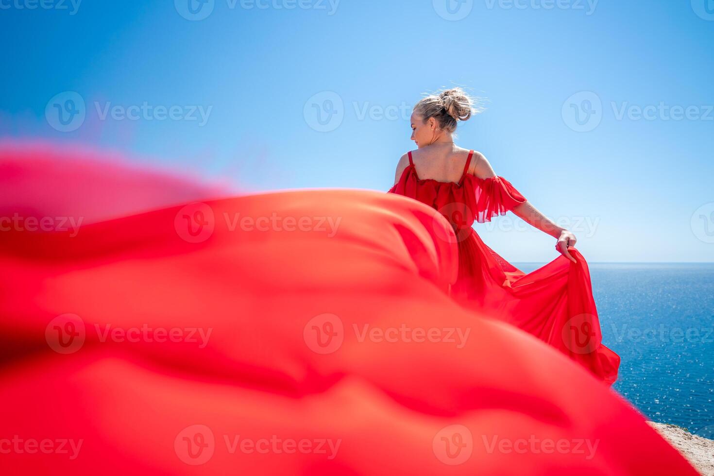 Blonde with long hair on a sunny seashore in a red flowing dress, back view, silk fabric waving in the wind. Against the backdrop of the blue sky and mountains on the seashore. photo
