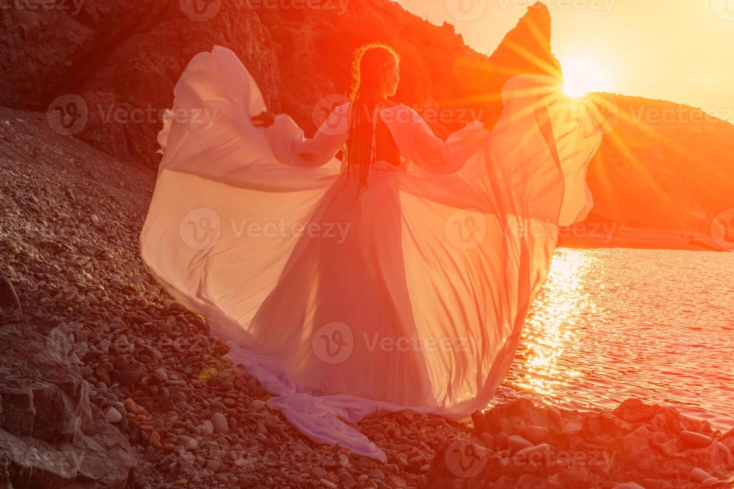 Mysterious woman silhouette long hair walks on the beach ocean water, sea nymph wind listens to the wave. Throws up a long white dress, a divine sunset. Artistic photo from the back without a face