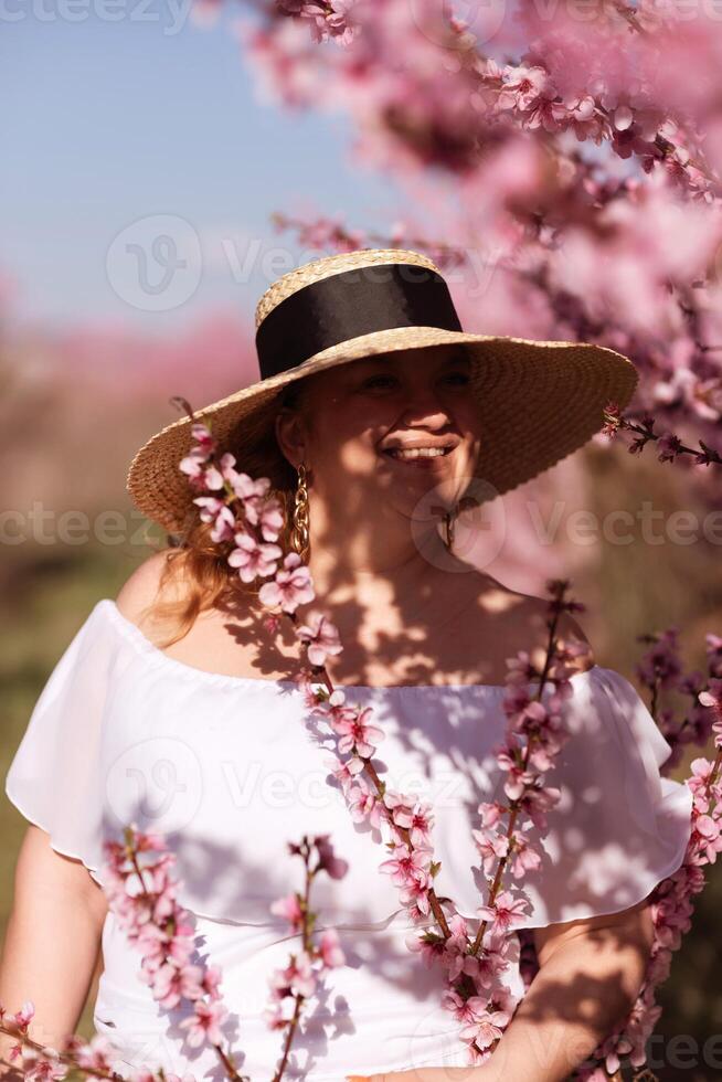 Woman blooming peach orchard. Against the backdrop of a picturesque peach orchard, a woman in a long white dress and hat enjoys a peaceful walk in the park, surrounded by the beauty of nature. photo
