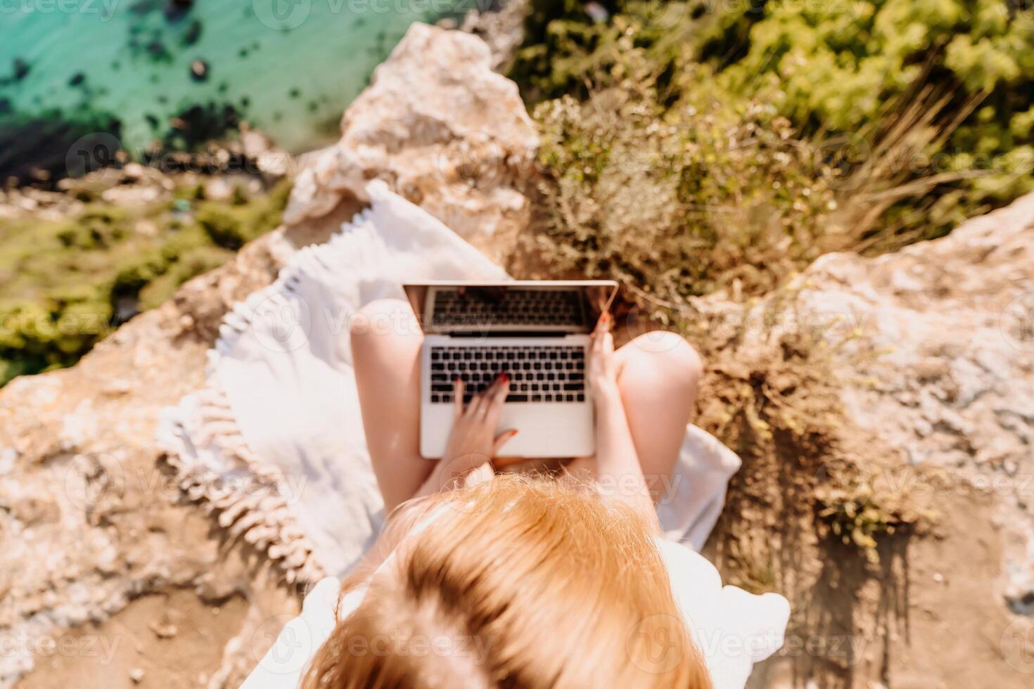 A woman is sitting on a rock overlooking the ocean with a laptop in front of her. She is enjoying the view and the peacefulness of the location. photo