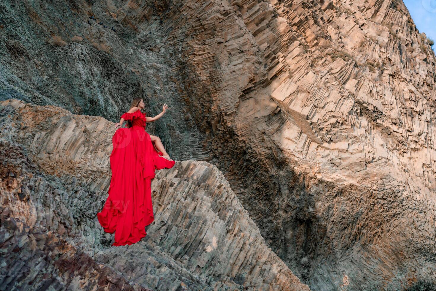 woman sea red dress. Woman with long hair on a sunny seashore in a red flowing dress, back view, silk fabric waving in the wind. Against the backdrop of the blue sky and mountains on the seashore. photo