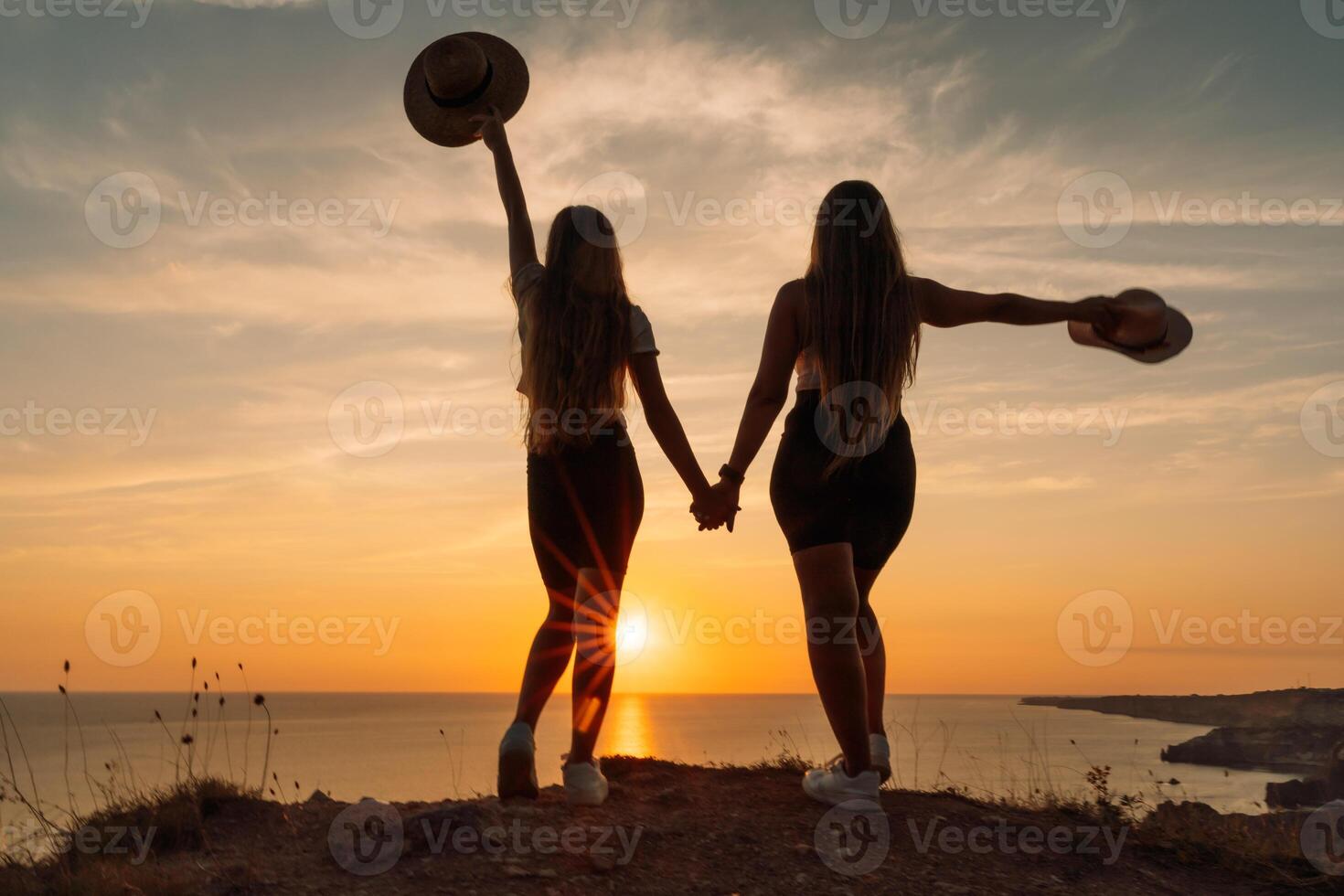 Two women are standing on a hillside, holding hands and wearing hats. The sun is setting in the background, creating a warm and peaceful atmosphere. photo