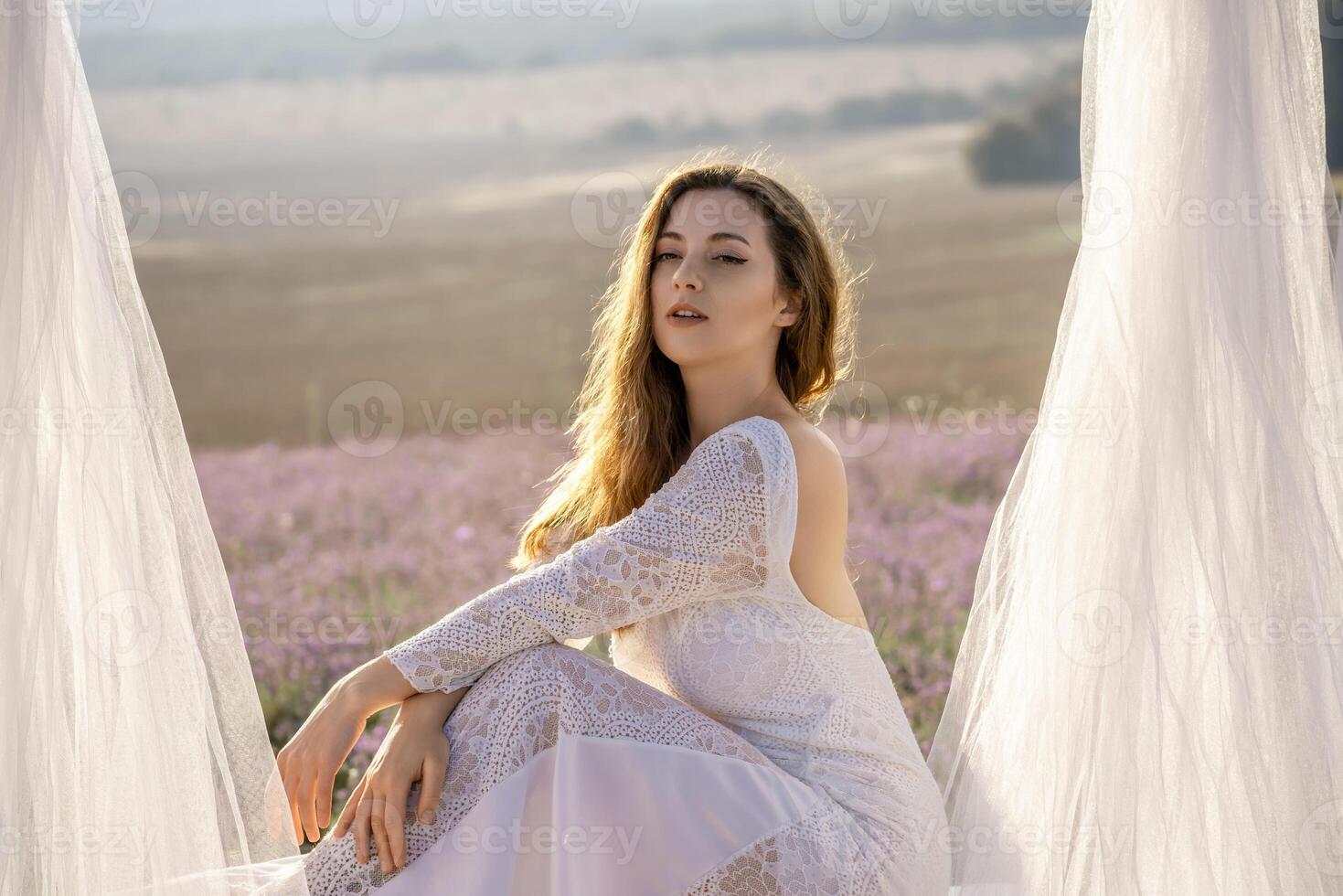 A woman in a white dress sits in a field with a white canopy above her. The scene is serene and peaceful, with the woman looking out over the landscape. photo