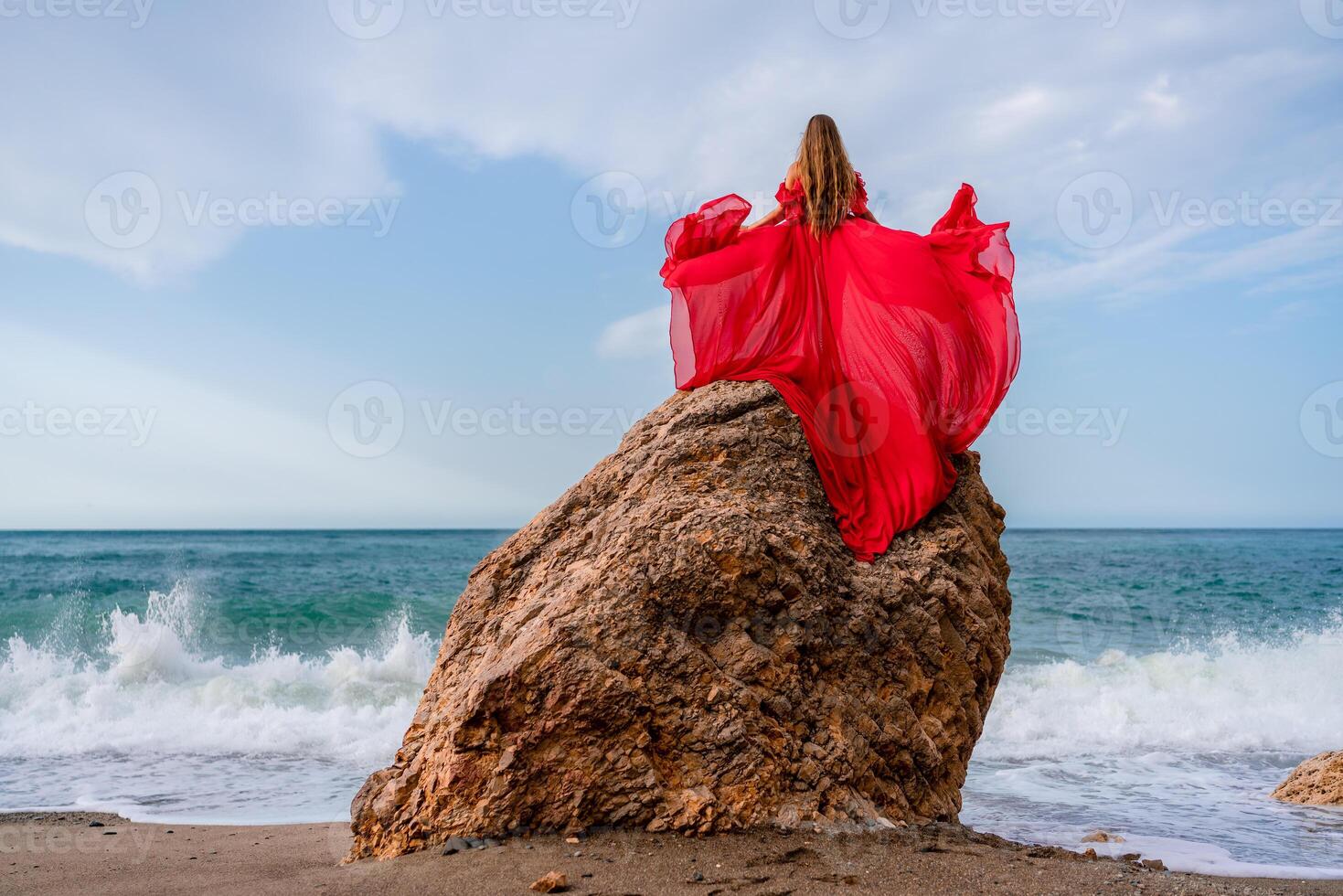 woman sea red dress. Woman with long hair on a sunny seashore in a red flowing dress, back view, silk fabric waving in the wind. Against the backdrop of the blue sky and mountains on the seashore. photo
