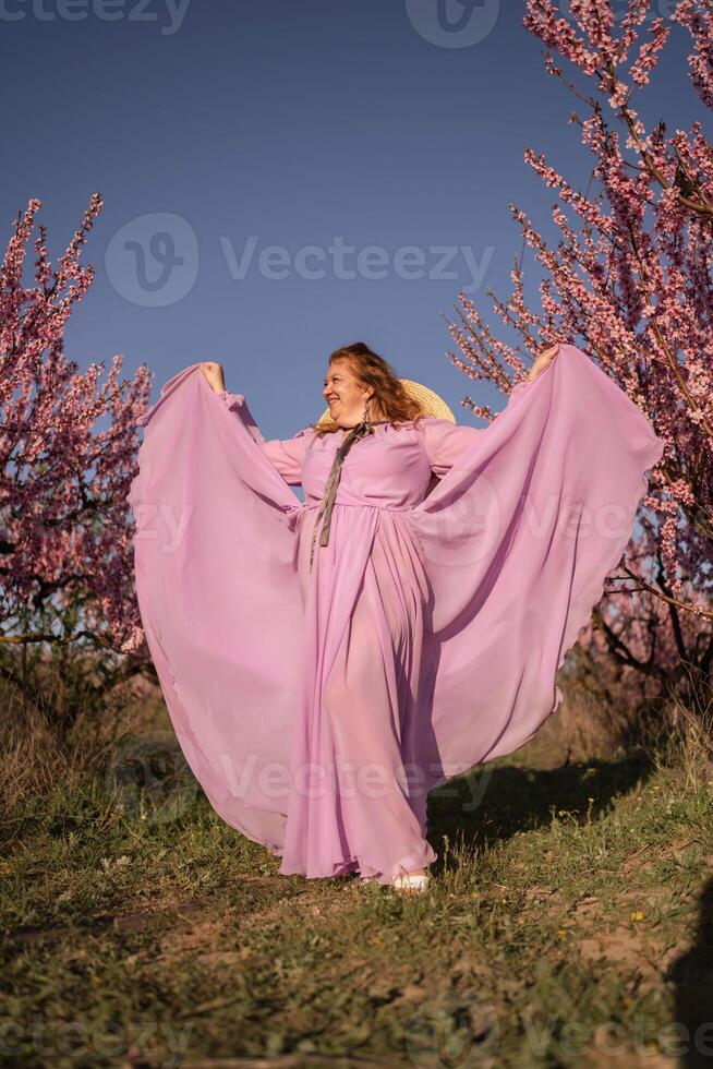 Woman blooming peach orchard. Against the backdrop of a picturesque peach orchard, a woman in a long pink dress and hat enjoys a peaceful walk in the park, surrounded by the beauty of nature. photo