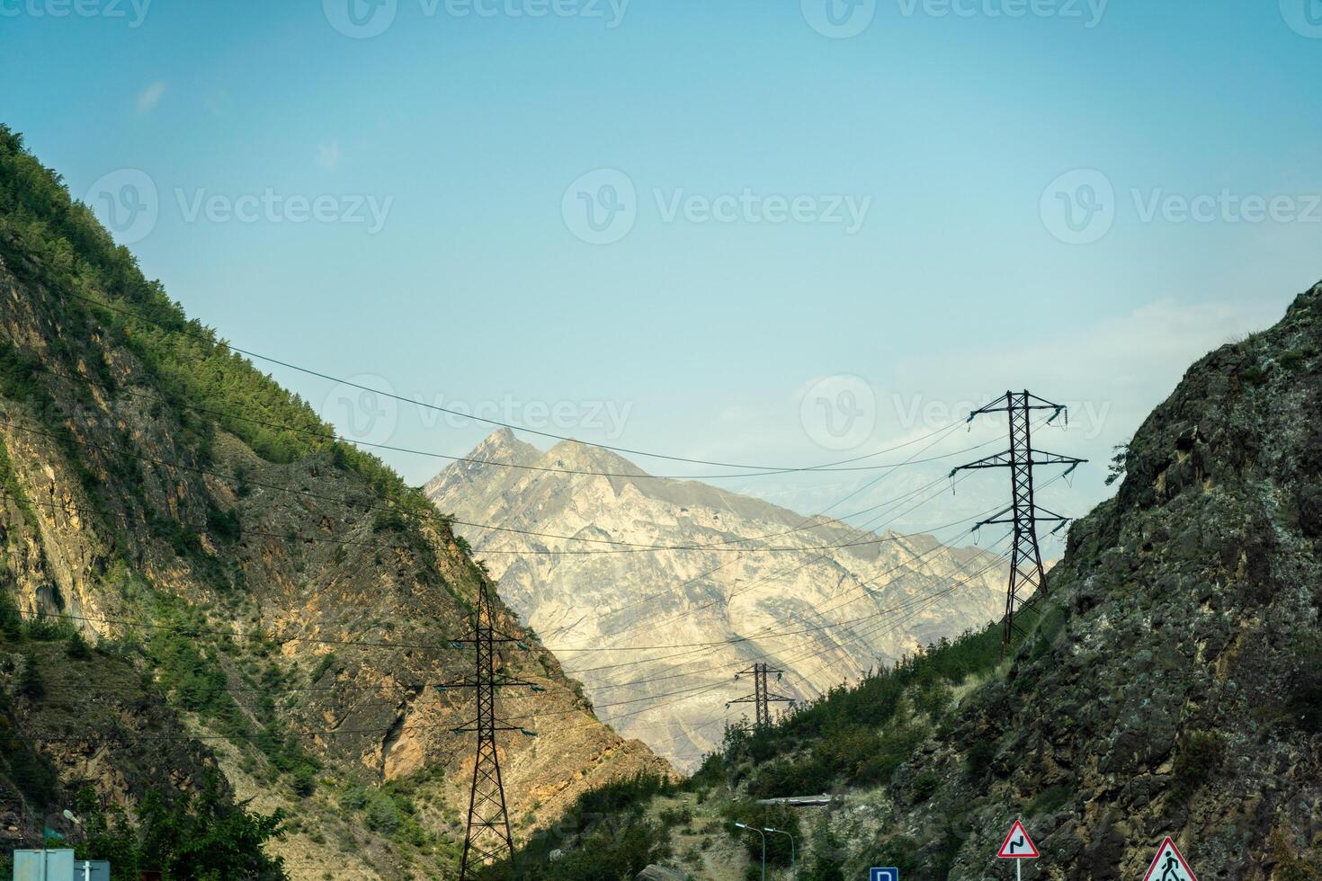 A mountain range with a power line running through it. The power line is high above the ground and is surrounded by trees. The mountains are covered in green foliage and the sky is clear and blue. photo